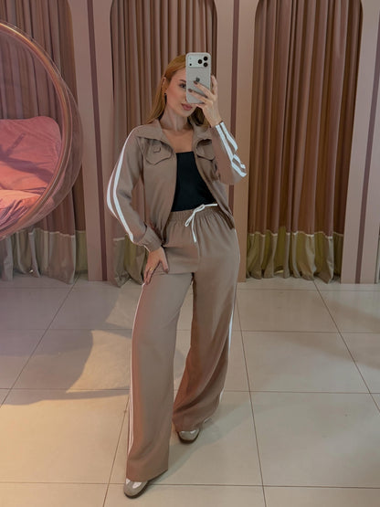 Modelo vestindo conjunto aerolook feminino bege com manga longa, jaqueta de zíper frontal e calça ampla com faixas laterais brancas.