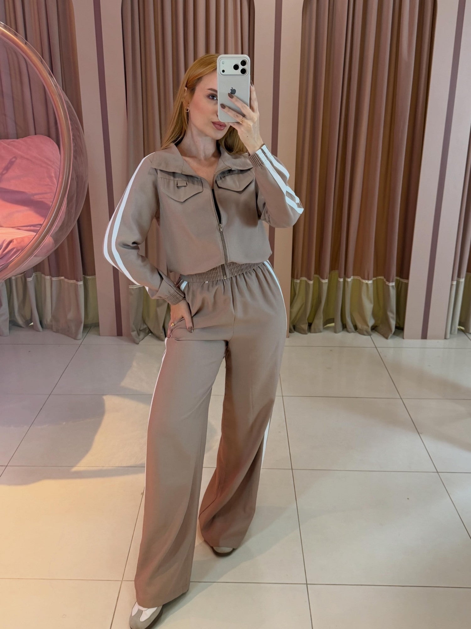 Modelo vestindo conjunto aerolook feminino bege com manga longa, jaqueta de zíper frontal e calça ampla com faixas laterais brancas.
